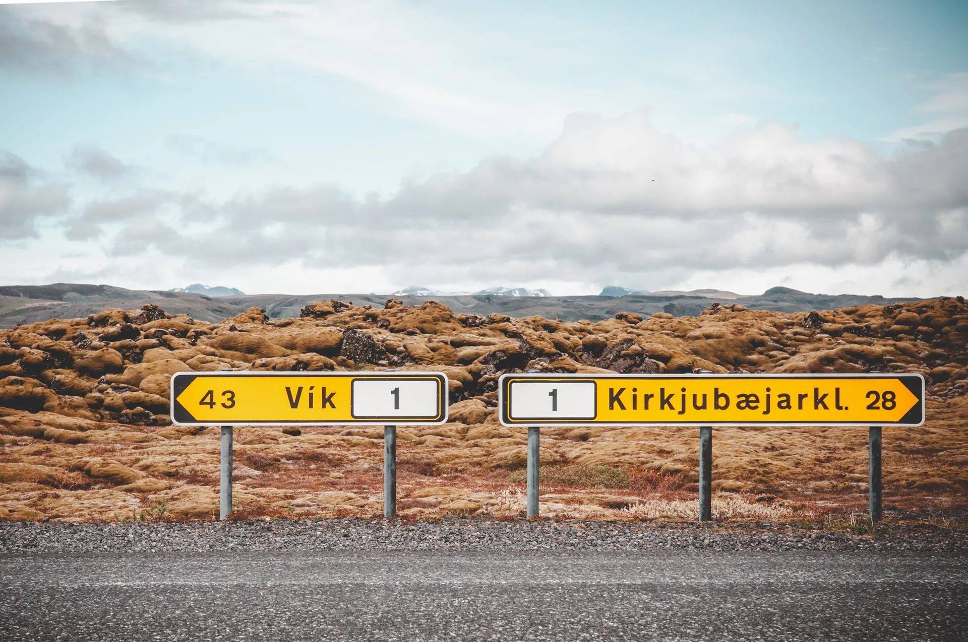 Two yellow Icelandic road signs at a Ring Road junction pointing to Vík and Kirkjubæjarklaustur across a golden moss-covered lava field, with snow-streaked mountains on the horizon under an overcast sky