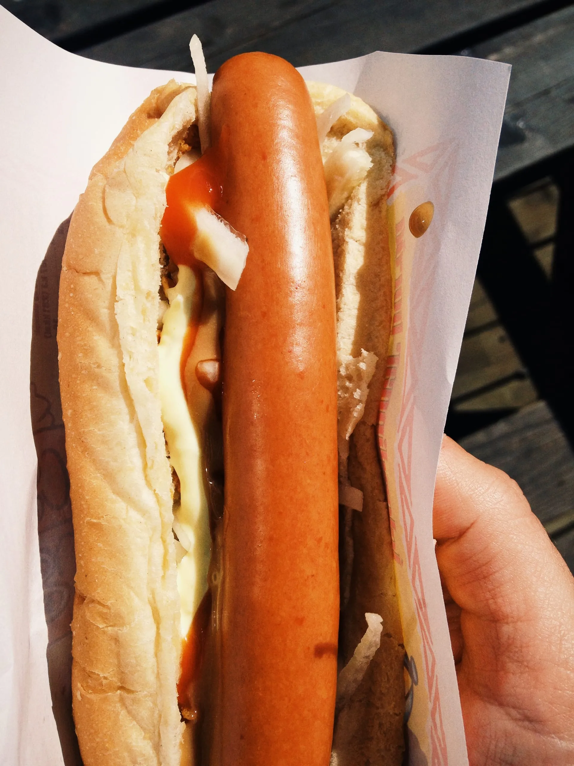 A hand holding a loaded Icelandic hot dog (pylsa) "með öllu" in its paper wrapper, the lamb-forward sausage in a steamed bun with ketchup, pale remoulade, brown mustard and raw and crispy fried onion, on a sunny day