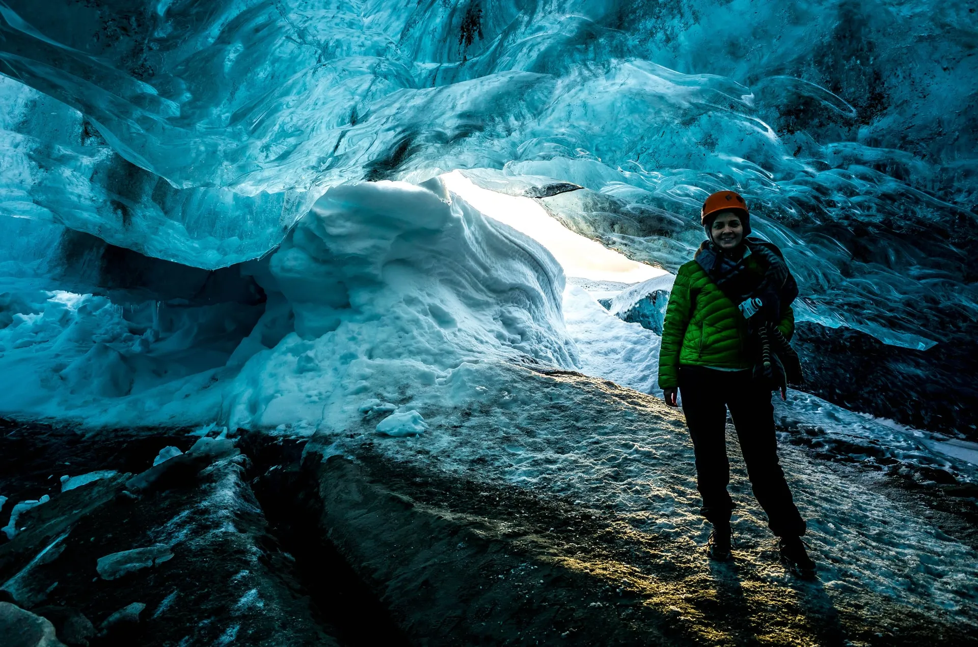 A visitor in an orange climbing helmet and crampons stands at the daylit mouth of a natural blue glacier ice cave in Southeast Iceland in January, with snow and low winter sun just beyond the scalloped ice roof