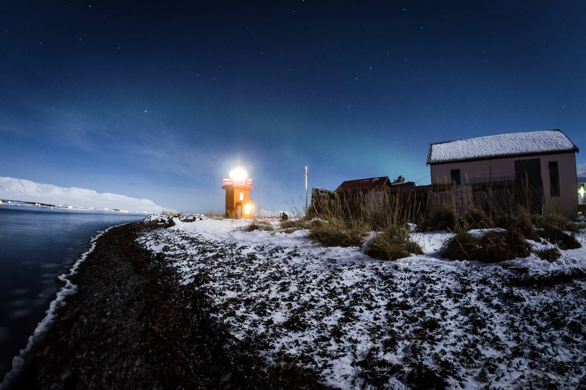 The northern lights rippling green over a dark fjord and a small lighthouse in North Iceland on a late-December night