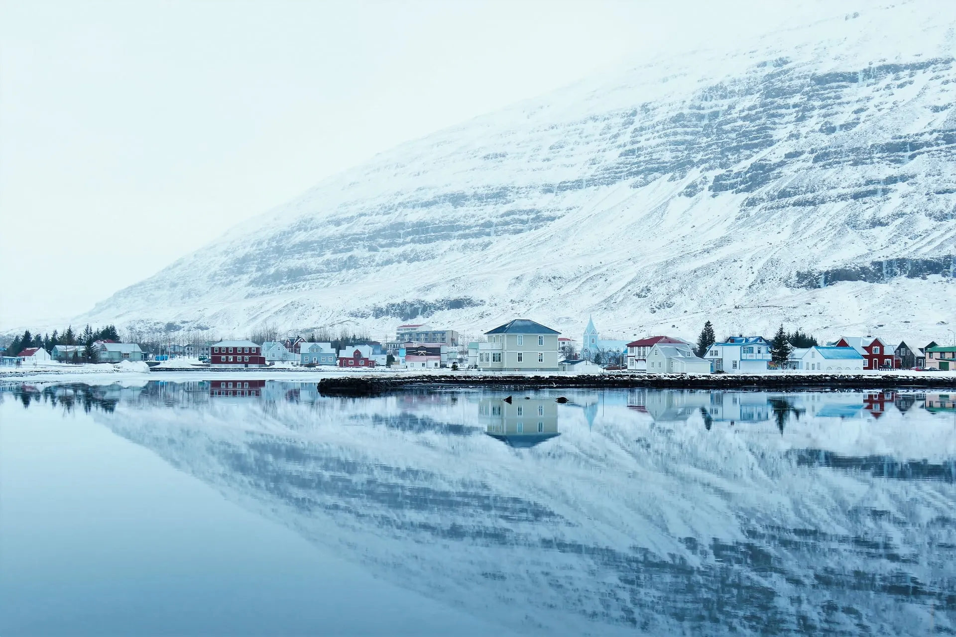 A snow-dusted mountain mirrored in the still water of an Icelandic fjord village under soft, low early-winter light