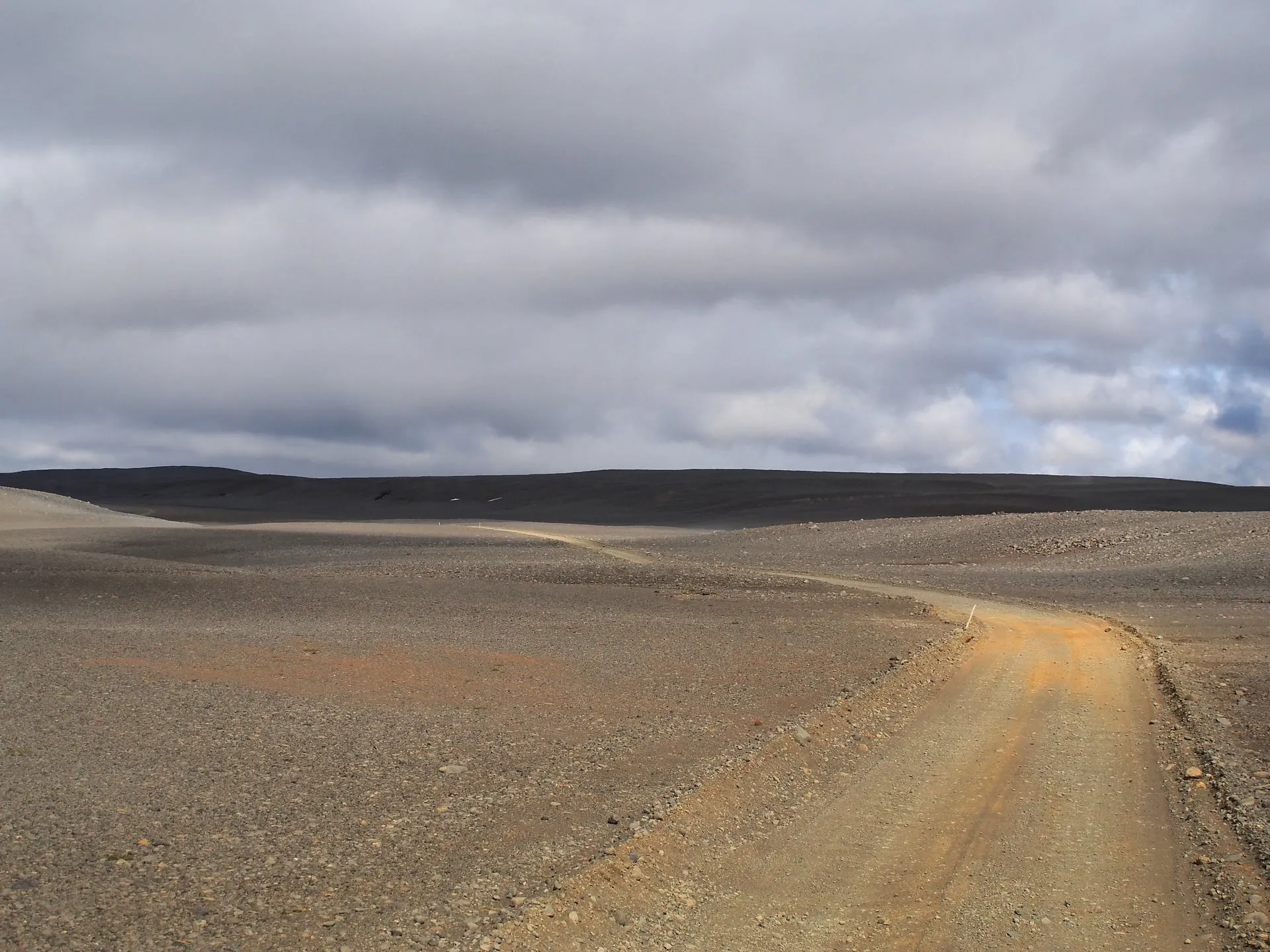 An empty gravel Highland F-road (Route F26 across Sprengisandur) curving through Iceland's grey volcanic desert interior toward low dark hills under a brooding overcast summer sky.
