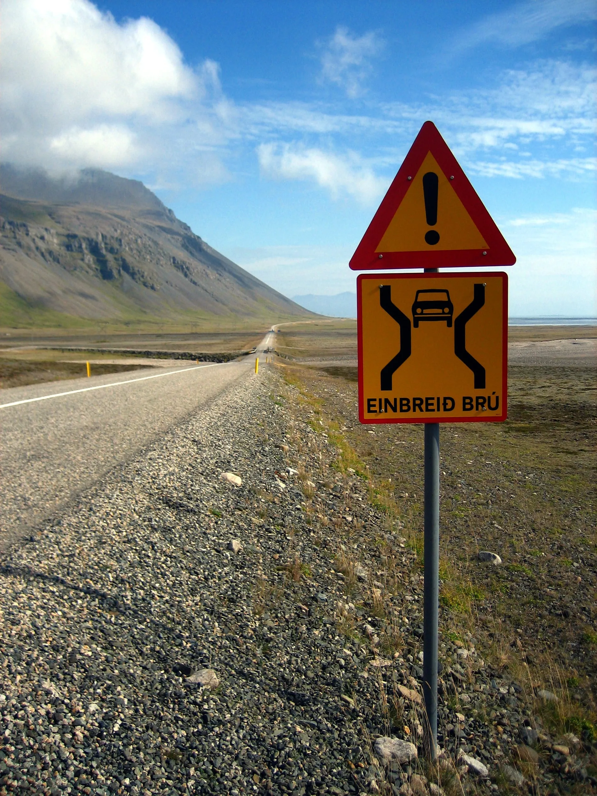 A yellow Icelandic einbreið brú warning sign, a red-bordered triangle above a pictogram of a car funnelling into a single lane, stands beside a paved road that narrows toward a one-lane bridge below a mountain and blue sky in East Iceland.