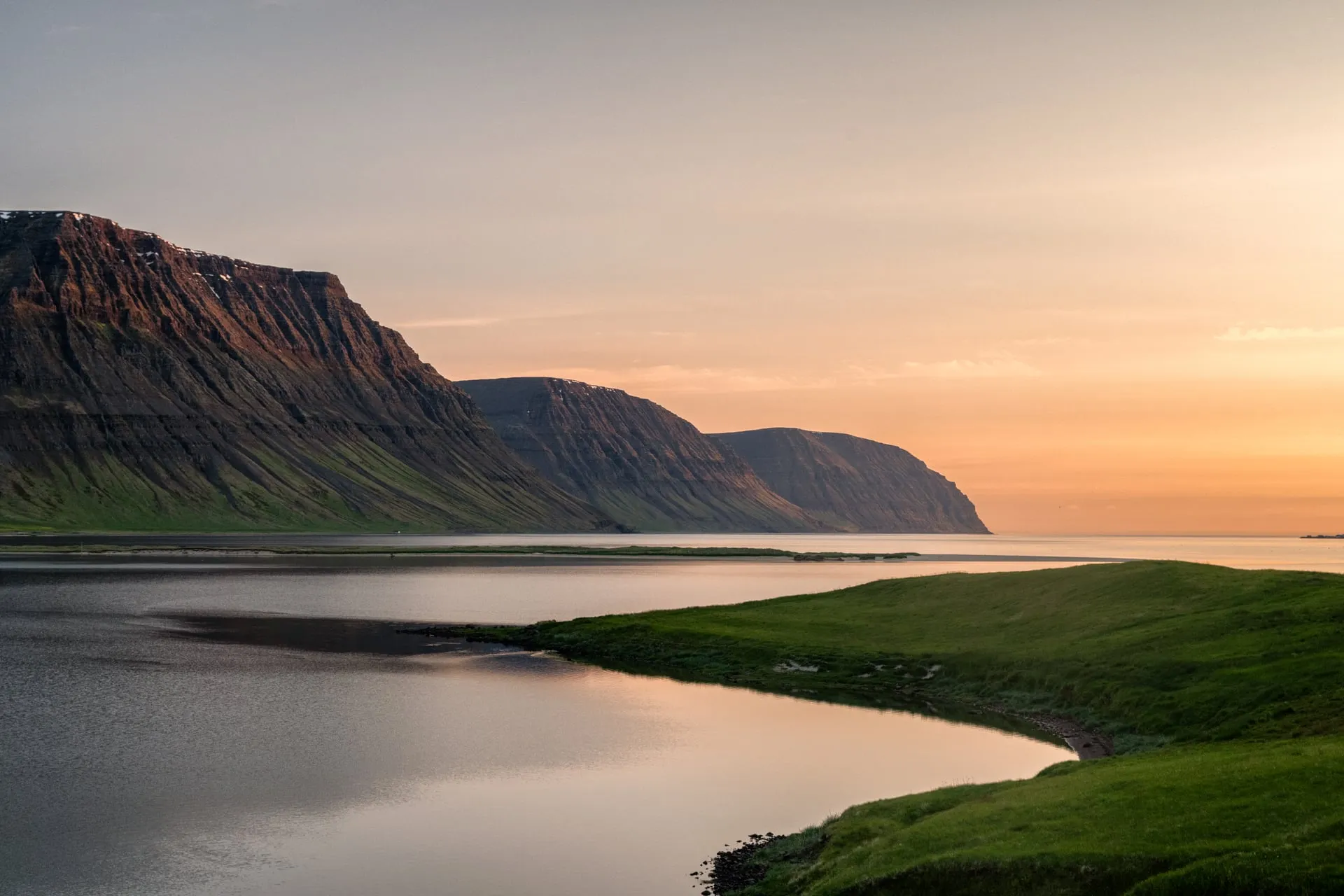 The midnight sun glowing low and golden along the horizon over a Westfjords coastline near Flateyri, the sky a long orange twilight reflected in calm water below green hills