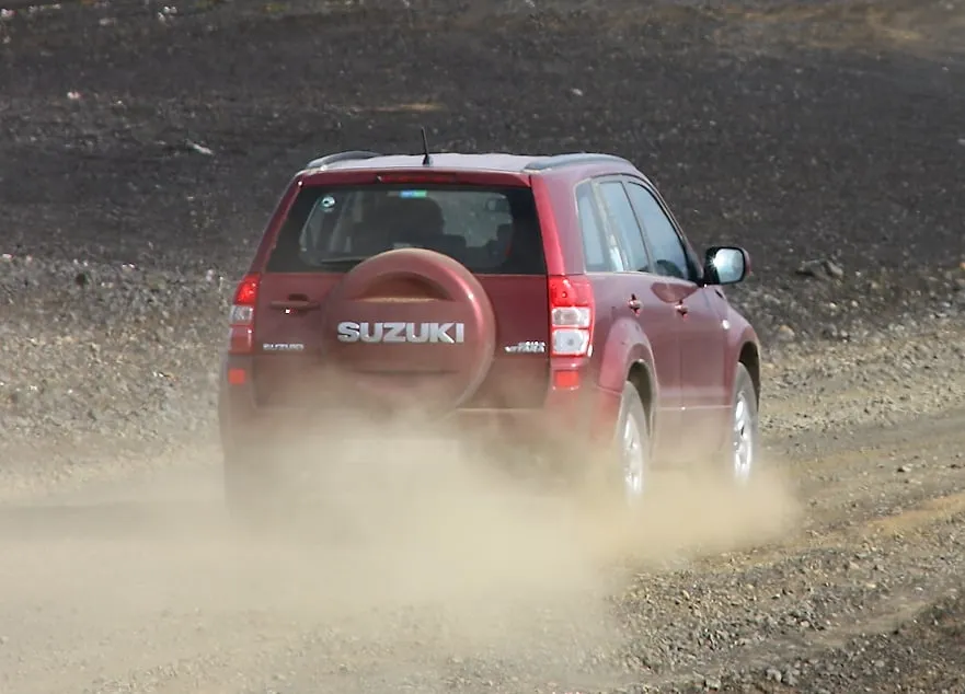 A dark-red Suzuki Grand Vitara 4x4 driving away on a loose grey gravel road across Iceland's interior, kicking up a plume of dust from its rear wheels.