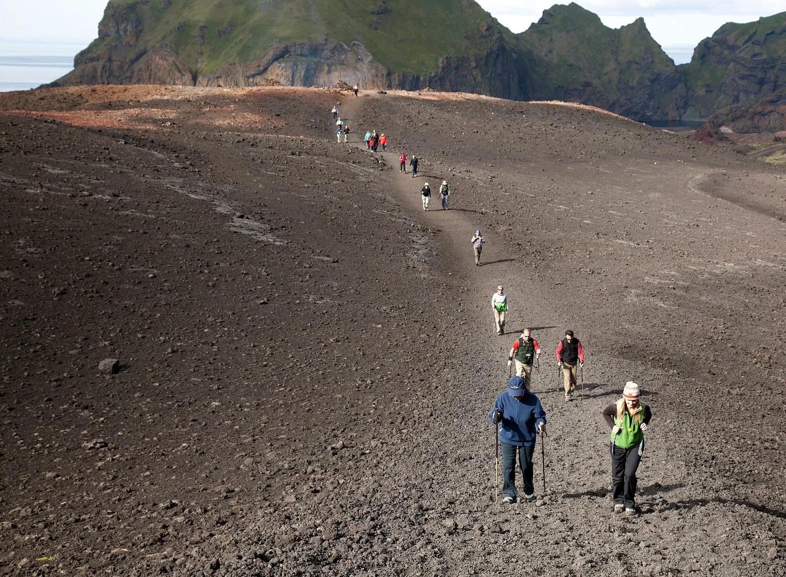 A line of hikers in layered outdoor clothing and waterproof wind jackets, two with trekking poles, climbing a volcanic gravel trail in Iceland on a bright summer day