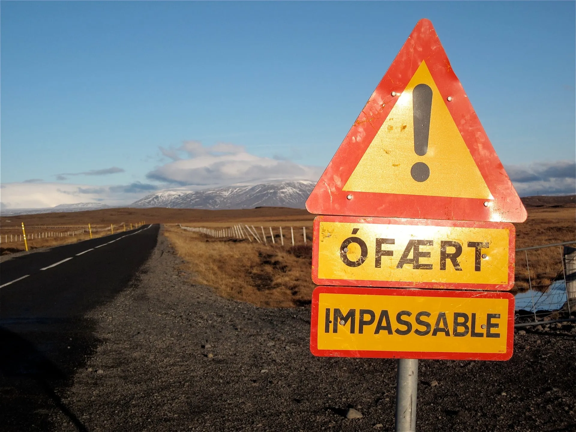 A weathered red-and-yellow Icelandic warning sign reading ÓFÆRT / IMPASSABLE at the start of the Kjölur Highland road just north of Gullfoss, the empty road running on across golden-brown late-autumn moorland toward snow-dusted mountains under a blue sky