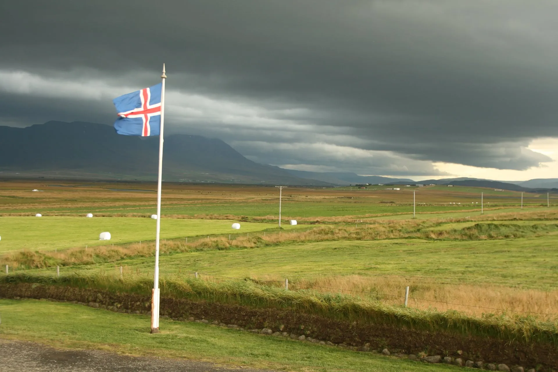 The Icelandic national flag streaming out horizontally from its pole in a stiff wind over green summer farmland, beneath a dark brooding storm sky with mountains behind in northwest Iceland