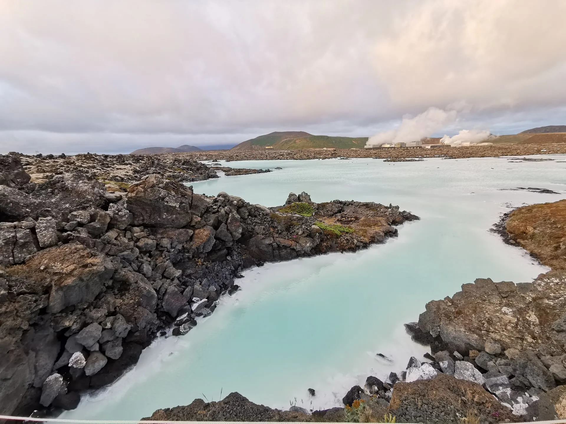 The Blue Lagoon's opaque milky-blue geothermal water pooling between crusted white silica and black Reykjanes lava rock, with steam rising from the Svartsengi geothermal plant on the horizon
