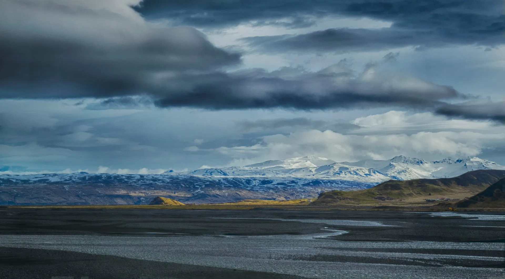 Heavy storm clouds sweeping low over dark Icelandic mountains and a glacial outwash plain, with shafts of light breaking through the changing weather